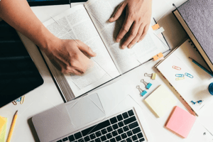 Manos trabajando en una investigación académica con libros, laptop y material de oficina, simbolizando el proceso de planificación y redacción eficiente descrito en el artículo del blog.